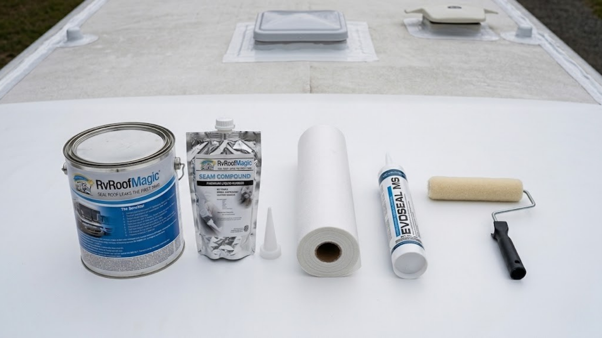 RV Roof Magic sealant can, seam compound, fabric roll, EVOSEAL MS caulk, and a paint roller arranged on a white RV roof.